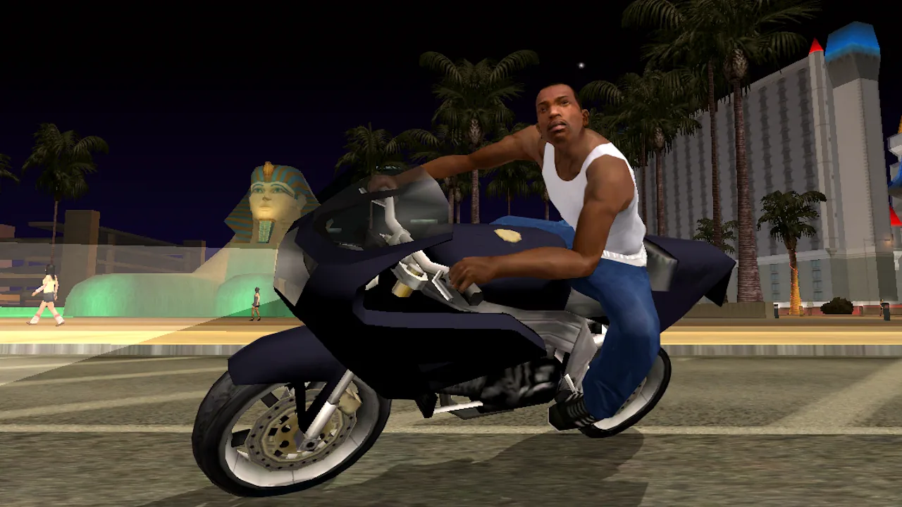 Captura de GTA: San Andreas com Carl Johnson numa mota desportiva escura, à noite, numa avenida com uma esfinge iluminada e um edifício alto de néon ao fundo